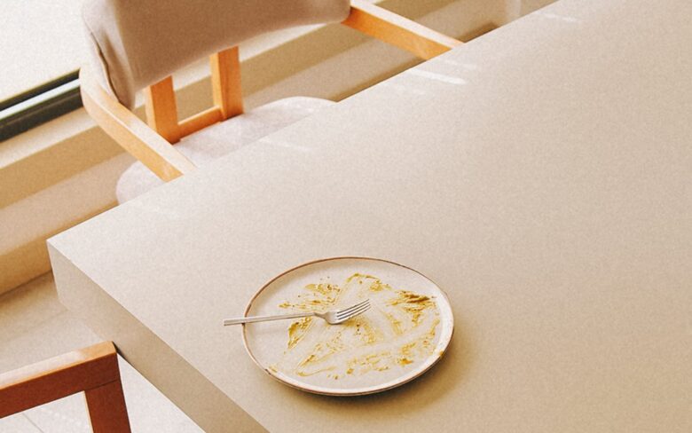 Pale dining table after a meal, a plate with leftovers and a fork, empty wooden chairs around it