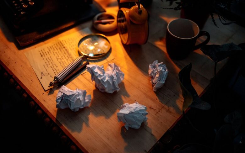 Desk at night with balls of crumpled paper, a magnifying glass over a handwritten page and a mug, in warm light